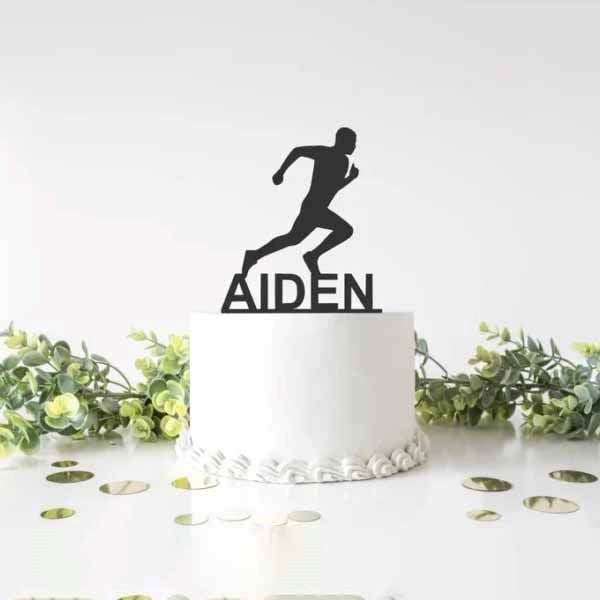 The image features a white round cake with a smooth surface. Atop the cake is a black acrylic cake topper depicting a running figure along with the name "AIDEN" in bold letters. Surrounding the cake are some green foliage and scattered circular gold confetti on the surface beneath it. The backdrop is a simple, light-colored setting that emphasizes the cake and decorations.