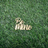 A gold, cursive cake topper that reads "Be Mine," positioned on a green artificial grass background.