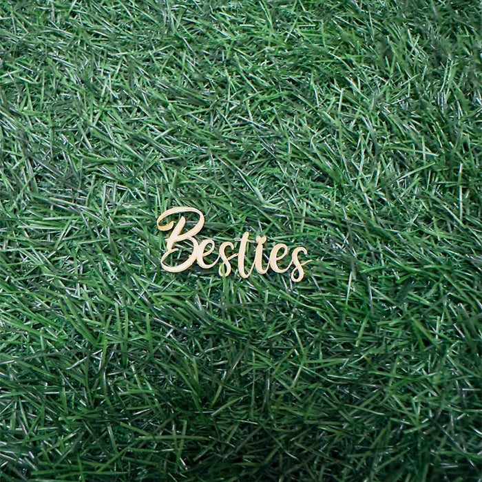 A wooden cake topper with the word "Besties" in a cursive font, lying on a green grass-like surface. The topper is light-colored and stands out against the background.