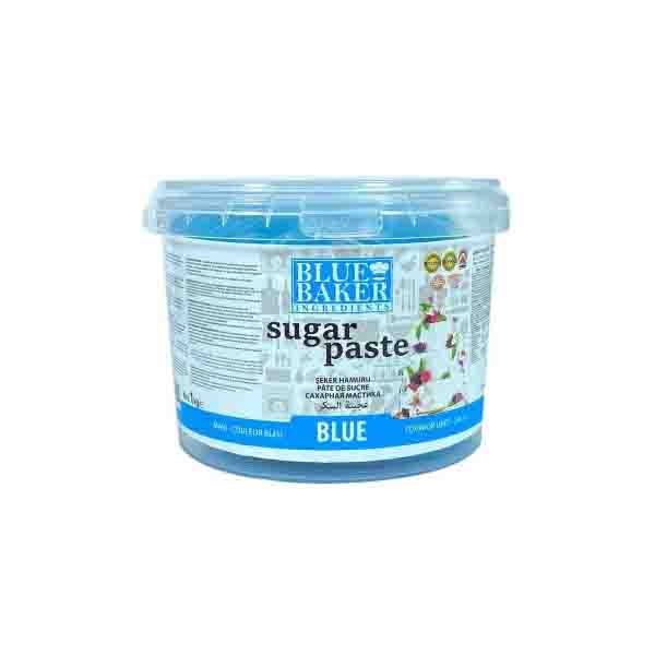 A clear plastic container of blue sugarpaste fondant is shown. The container features a label with the brand name "Blue Baker" at the top, prominently displaying the words "sugar paste" in bold lettering. Below, the color designation "BLUE" is highlighted in a blue band. The container is sealed with a transparent lid, and additional text and images of cake decorations are present on the label, suggesting its use for baking and decorating cakes.