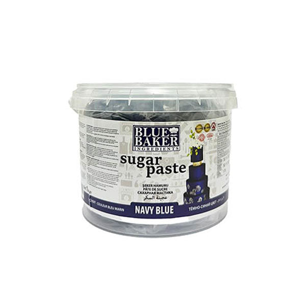 A clear plastic tub containing navy blue sugar paste labeled "Blue Baker Ingredients" is shown. The tub has a white lid and features the product name "sugar paste" prominently on the front. Additional details include the word "NAVY BLUE" at the bottom. The background displays decorative graphics associated with baking and cake decorating. The container is 1 kg in size.