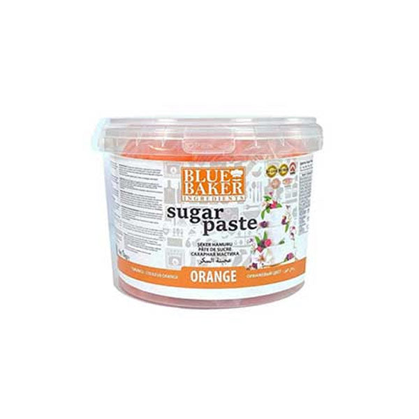 A clear plastic container of orange sugar paste fondant labeled "Blue Baker." The container has a white lid and features a colorful design with the product name, "sugar paste," and "ORANGE" prominently displayed on the front. The packaging includes instructions and additional information. The background shows decorative elements, suggesting baking or cake decoration use.