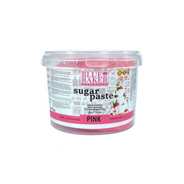A round, clear plastic container of pink sugar paste labeled "Blue Baker Sugar Paste" in bold black text. The container holds 1 kg of product, and features decorative images of a cake with flowers. The label includes additional information and icons indicating its use. The container has a pink band at the bottom with the word "PINK."