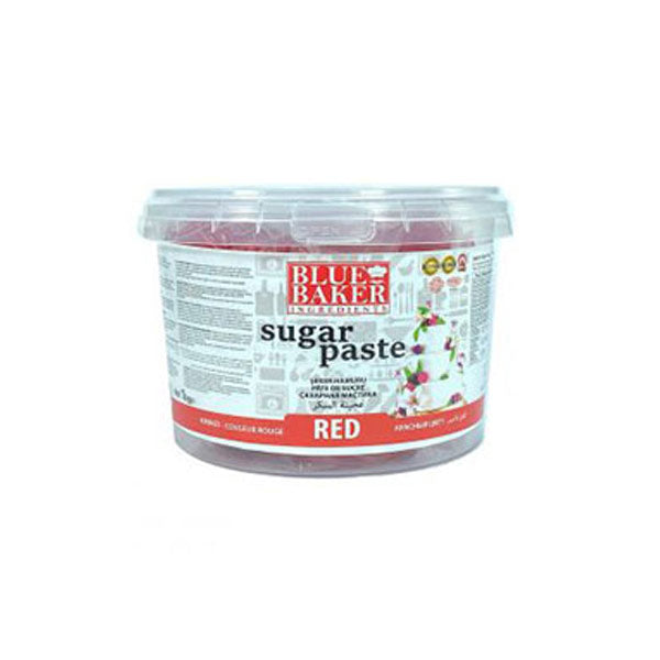The image shows a plastic container of Blue Baker red sugar paste, weighing 1 kilogram. The container is transparent with a label on the front that features the brand name "Blue Baker" prominently at the top. Below the brand name, the words "sugar paste" are displayed, followed by the word "RED" in bold red letters at the bottom. The background of the label includes various cake decorating icons and information. The lid of the container is clear, revealing the red sugar paste inside.