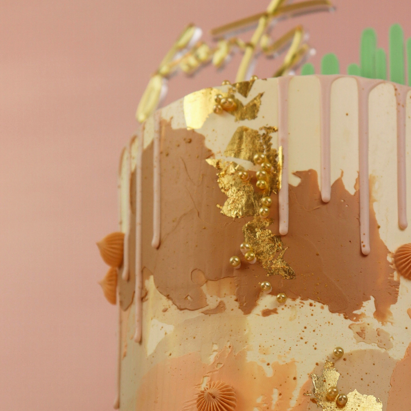 The image features a decorated cake with a smooth surface showcasing a blend of pastel colors, primarily peach and cream. It has vertical drips of a lighter color at the top and is adorned with gold leaf accents, edible pearls, and decorative elements such as ribbed fondant shapes. The background is a soft pink, enhancing the overall aesthetic of the cake. A festive cake topper is positioned near the top, complementing the elegant design.
