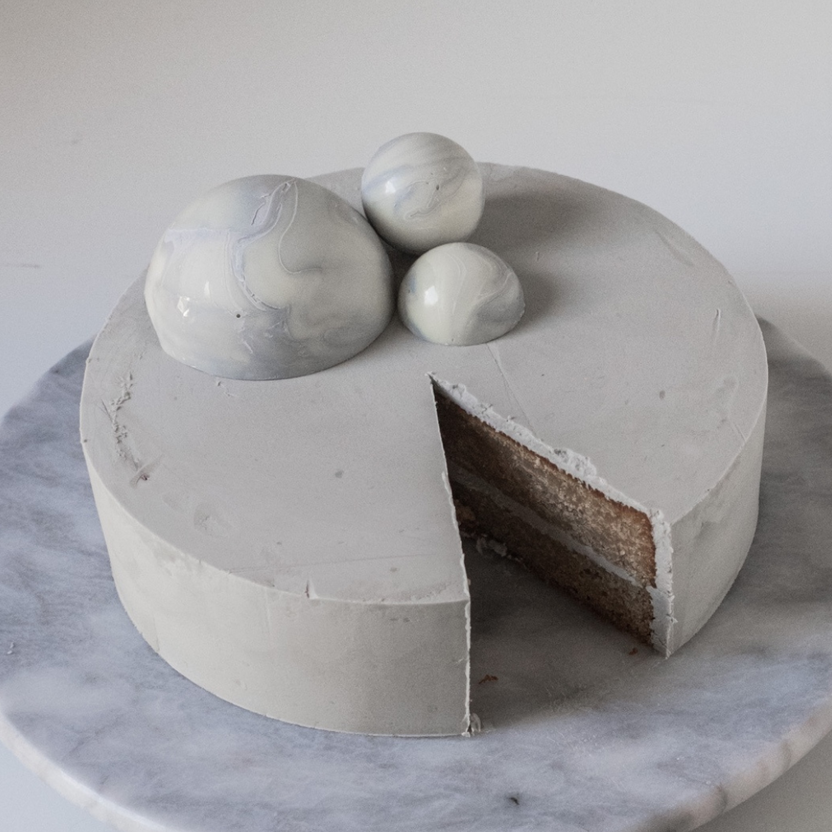 A round cake with a smooth, gray frosting sits on a marble cake stand. The cake has a slice removed, revealing layered sponge and filling inside. Three decorative spheres, resembling marbled stone, are positioned on top of the cake, adding a modern aesthetic. The background is simple and light, emphasizing the cake&