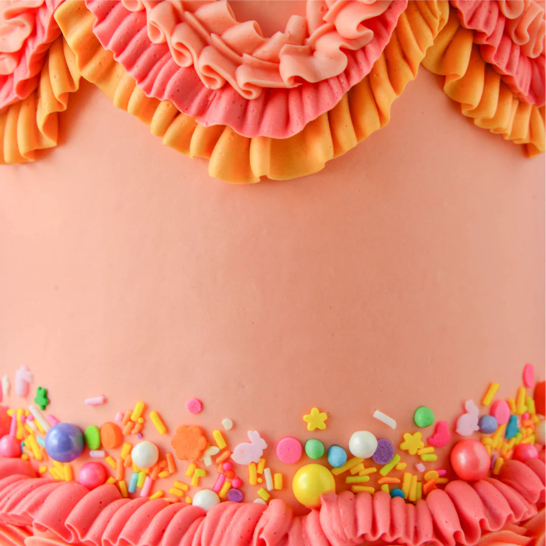 The image features a close-up of a decorated cake. The cake has a smooth coral-colored surface adorned with ruffled icing in varying shades of pink and orange at the top. At the base, there is a colorful array of sprinkles, including small stars, circles, and various shapes, in shades of yellow, pink, green, blue, and purple. The overall appearance is vibrant and festive, highlighting the cake&