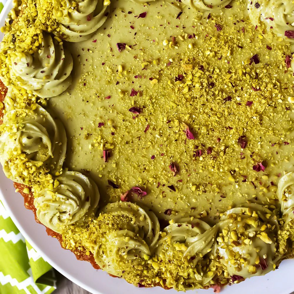 Close-up of a vibrant pistachio tart with creamy green swirls and a generous sprinkle of crushed pistachios and rose petals, set on a white plate.
