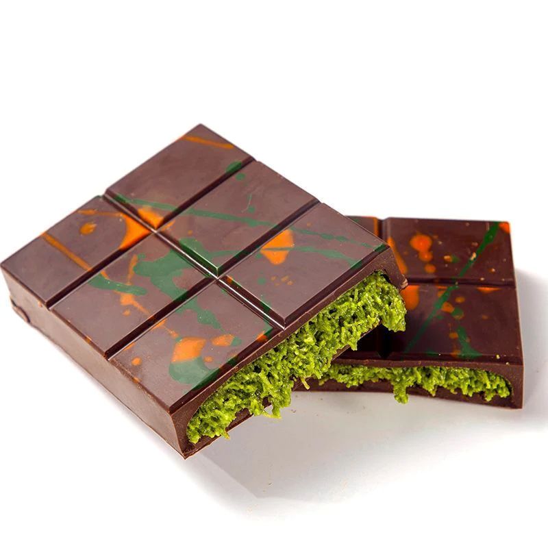 A chocolate bar with a green filling is decorated with splashes of orange and green. One piece is broken, revealing the textured filling inside.