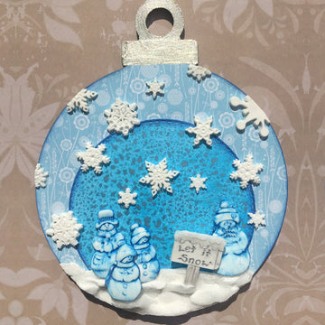 A round, festive ornament featuring a winter scene. The background is light blue with a textured pattern. In the center, three snowmen are gathered around a sign that reads "Let it Snow." Snowflakes are scattered throughout the design, with additional snowflakes and a white, snowy ground detail at the bottom. The ornament has a silver hanger at the top.
