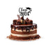 A layered chocolate cake topped with a glossy chocolate glaze and decorated with chocolate curls and swirls. Atop the cake, there is an acrylic topper that reads "Class of 2024" in black font, accompanied by a graduation cap graphic. The cake sits on a white background.