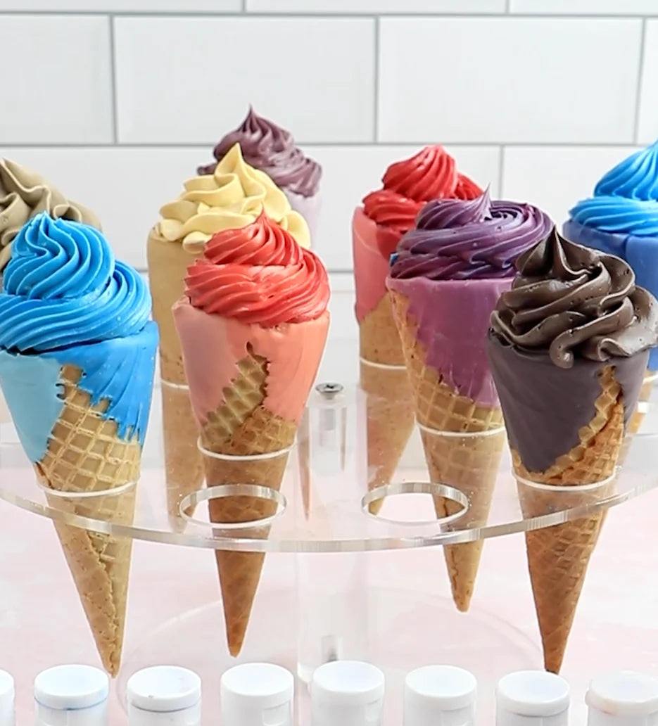 The image features a display of several ice cream cones arranged on a clear stand. Each cone is topped with swirls of colorful frosting, resembling different ice cream flavors. The colors include blue, pink, orange, yellow, red, purple, and brown. The cones are made of a traditional waffle texture. In the foreground, there are small white containers, suggesting the presence of food coloring or similar products, with a light pink surface beneath. The background appears to have a tiled pattern.