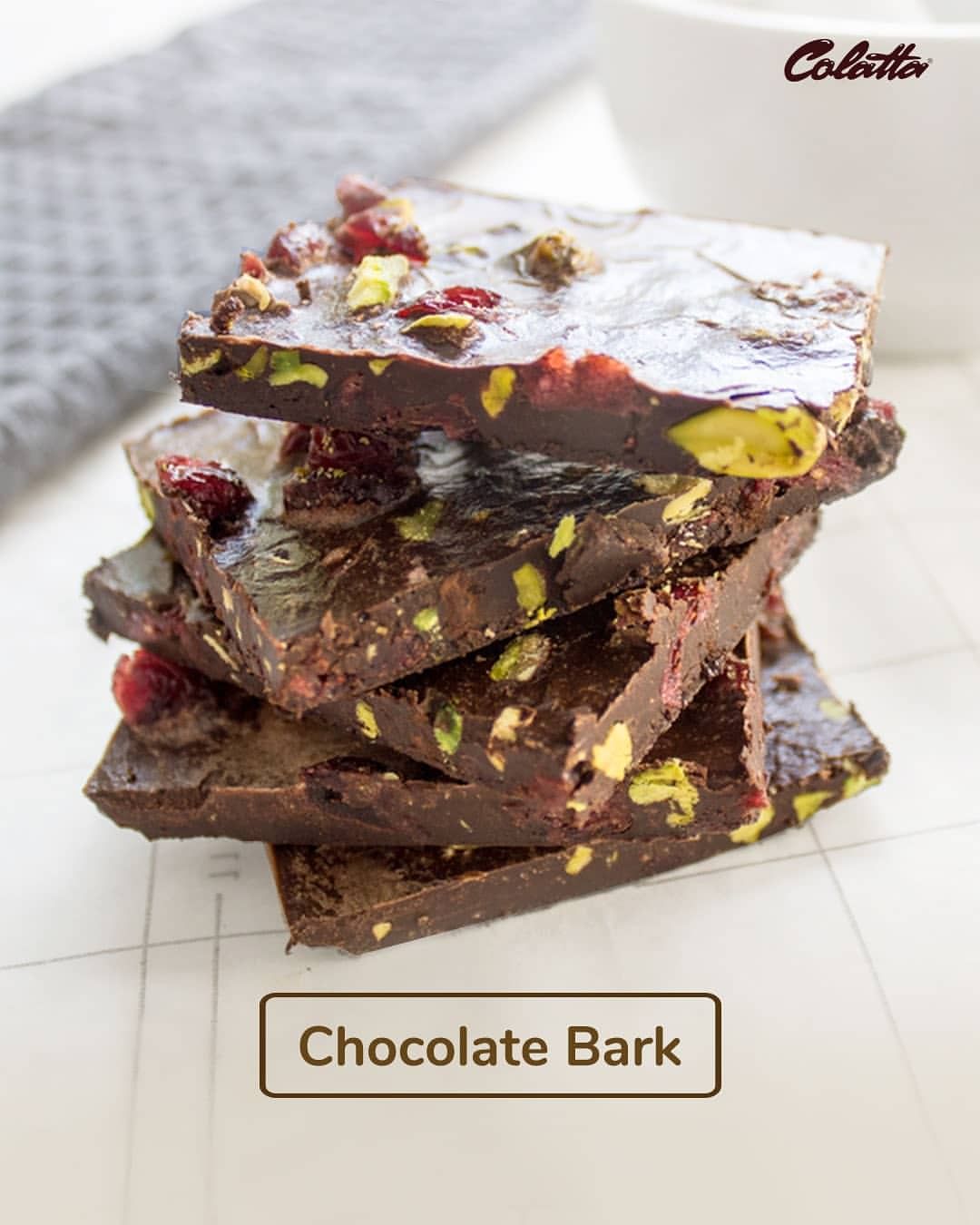 A stack of chocolate bark pieces, featuring dark chocolate mixed with nuts and dried fruits, such as pistachios and cranberries. The background is a light surface with a grey textured cloth and a bowl visible. The text "Chocolate Bark" is displayed below the stack.