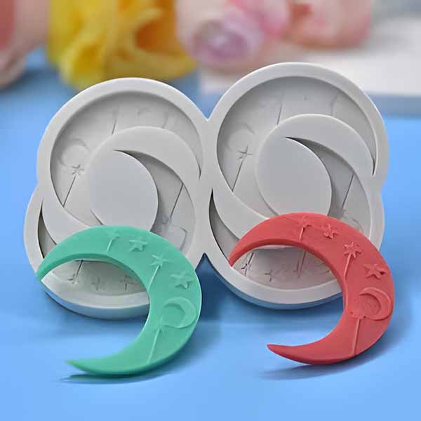 A 4-cavity silicone mold shaped like crescent moons is displayed against a light blue background. The mold is white and features four crescent moon impressions. In front of the mold, there are two finished moon-shaped pieces: one is a light green color and the other is red, with decorative star and moon designs.