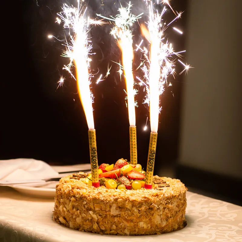 A round cake topped with fruit, including strawberries and yellow fruits, is displayed on a table. Three sparkler candles are positioned vertically in the cake, emitting bright sparks and flames against a dark background. The cake is garnished with nuts and has a textured surface, while a white plate and cutlery are visible nearby.