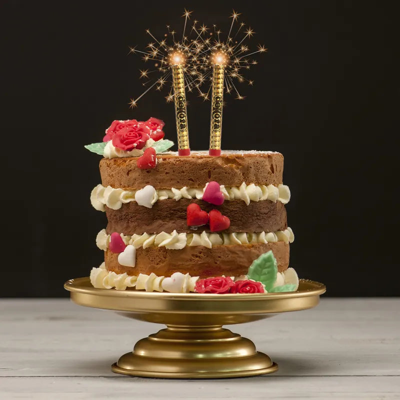 A three-layered cake is displayed on a gold cake stand. The cake features alternating layers of light and dark sponge, decorated with white frosting. Colorful fondant decorations, including red roses and heart shapes in pink and white, embellish the cake. At the top, two golden sparkler candles are lit, emitting sparks and creating a festive atmosphere, set against a dark background.