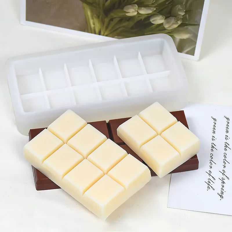 A silicone chocolate bar mold is displayed alongside two chocolate bars made from the mold. The mold is white and rectangular, featuring eight sections for shaping the bars. One bar is brown, resembling traditional chocolate, while the other is cream-colored, shaped like a chocolate bar with eight segments. A small decorative card with a quote about color is partially visible in the background, along with a blurred image of flowers.