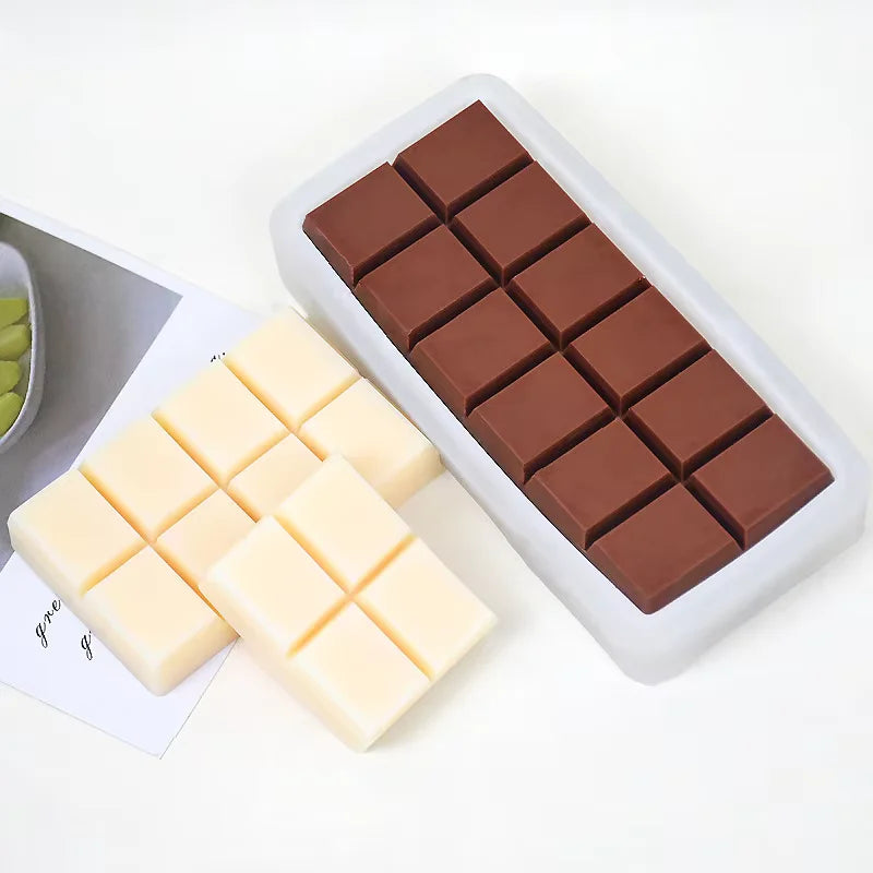 Image description for accessibility: The image features a silicone mold designed for making chocolate bars. In the center, there is a white silicone mold containing several dark chocolate squares that are arranged in a grid pattern. To the left, there are two pieces of white chocolate, also shaped like chocolate squares, positioned on a light background. The overall setting appears clean and minimalist.