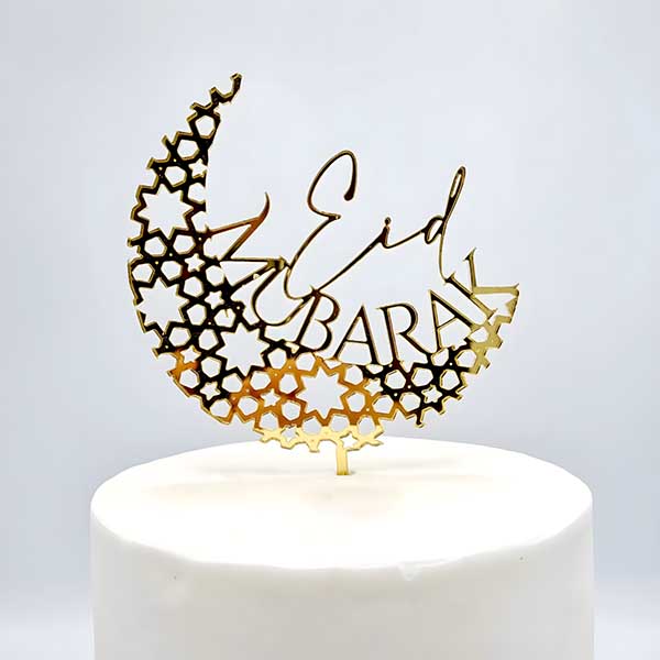 An elegant acrylic cake topper featuring a crescent moon design adorned with geometric star patterns, with the words "Eid Mubarak" artistically displayed. The topper is gold in color and is placed atop a white, round cake. The background is plain, enhancing the focus on the decorative piece.