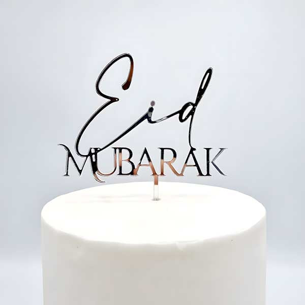 A shiny acrylic cake topper displaying the words "Eid Mubarak" in an elegant font. The topper is placed on top of a white cake, which has a smooth surface. The background is light and neutral, emphasizing the decorative piece.
