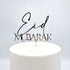 A shiny acrylic cake topper displaying the words "Eid Mubarak" in an elegant font. The topper is placed on top of a white cake, which has a smooth surface. The background is light and neutral, emphasizing the decorative piece.