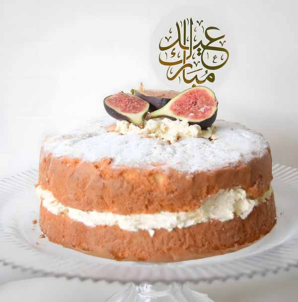 A round cake on a glass platter, layered with cream and topped with powdered sugar. The cake is decorated with fresh figs cut in half. Atop the cake is a decorative cake topper with Arabic calligraphy that reads "Eid Mubarak." The background is plain white, highlighting the cake&