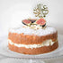 A round cake on a glass platter, layered with cream and topped with powdered sugar. The cake is decorated with fresh figs cut in half. Atop the cake is a decorative cake topper with Arabic calligraphy that reads "Eid Mubarak." The background is plain white, highlighting the cake&