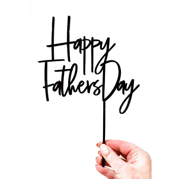 A hand is holding a black acrylic cake topper that reads "Happy Fathers Day" in a stylish script font. The topper features a thin stick for inserting into a cake, and it is displayed against a white background.