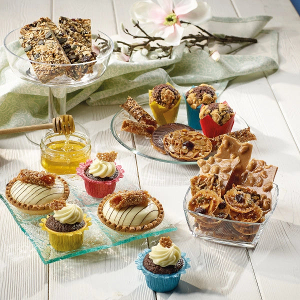 A variety of desserts on white wood: granola bars, cupcakes with creamy frosting, brittle, and honey in a jar. Floral decor adds elegance.