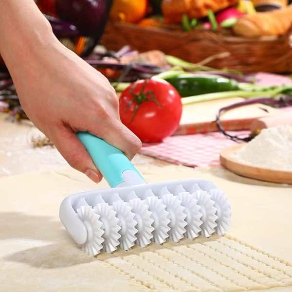 A hand is holding a fondant ribbon cutter, which features a textured rolling head with patterned cutters. The tool is being rolled across a flat surface of fondant, creating evenly spaced lines. In the background, there are various colorful vegetables and fruits, suggesting a vibrant kitchen setting.
