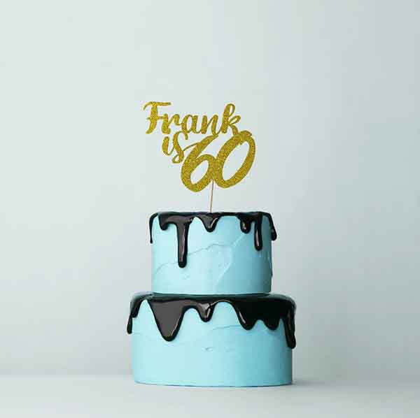 A two-tiered birthday cake with light blue frosting. The top tier has a glossy black drip effect along the edge. Atop the cake, there is a glittery gold cake topper reading "Frank is 60." The background is plain and pale, emphasizing the cake and topper.