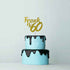 A two-tiered birthday cake with light blue frosting. The top tier has a glossy black drip effect along the edge. Atop the cake, there is a glittery gold cake topper reading "Frank is 60." The background is plain and pale, emphasizing the cake and topper.