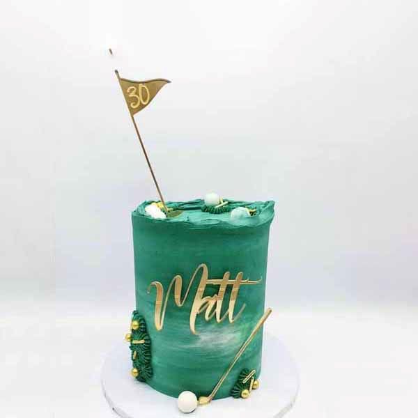 A round cake decorated in green frosting, featuring a gold flag with the number "30" on top. The name "Matt" is elegantly written in gold lettering on the front of the cake. Additional decorative elements include white and gold accents, resembling golf balls and a putting green. The cake is displayed on a white platter with a plain white background.