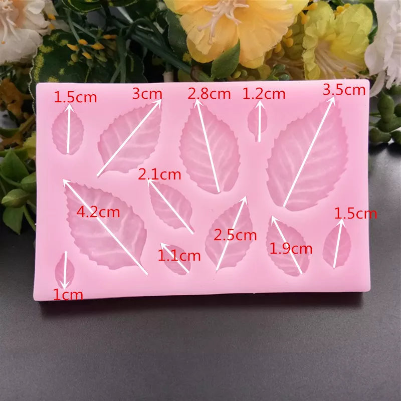 A pink silicone mould featuring various sizes of rose leaf shapes. The mould contains multiple leaf designs, each with measurements indicated in centimeters next to them. The background includes some decorative greenery and flowers, enhancing the presentation of the mould.
