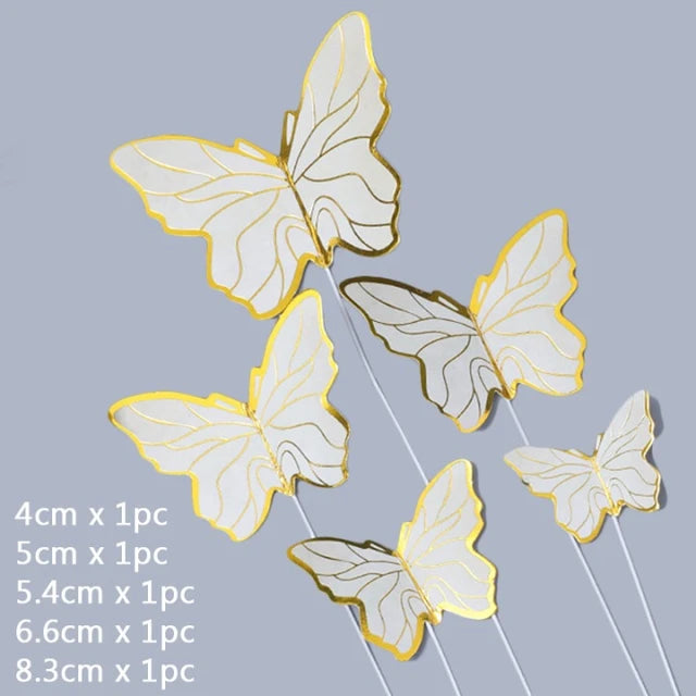 A collection of five decorative butterfly cake toppers, featuring a white and gold design. The sizes of the butterflies are labeled as 4 cm, 5 cm, 5.4 cm, 6.6 cm, and 8.3 cm. Each butterfly is attached to a thin stick for easy placement on a cake or dessert. The background is a plain gray color.