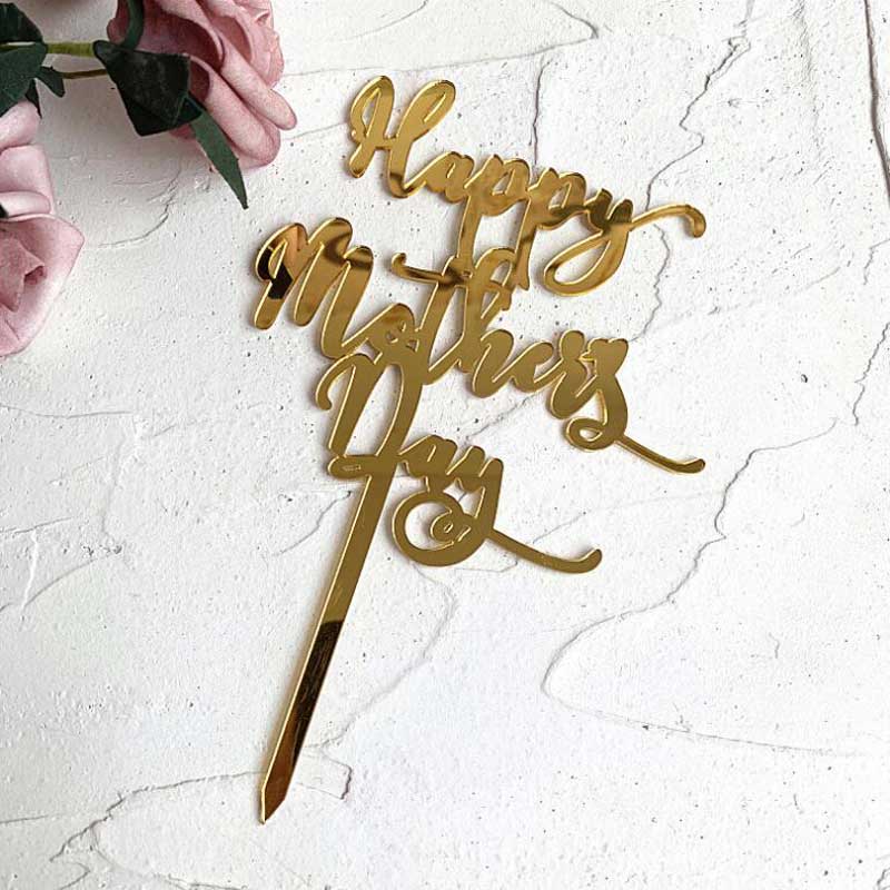 A gold acrylic cake topper with the inscription "Happy Mothers Day" in an elegant cursive font. The topper features a stake for insertion into a cake and is placed on a textured white surface, accompanied by soft pink roses in the background.