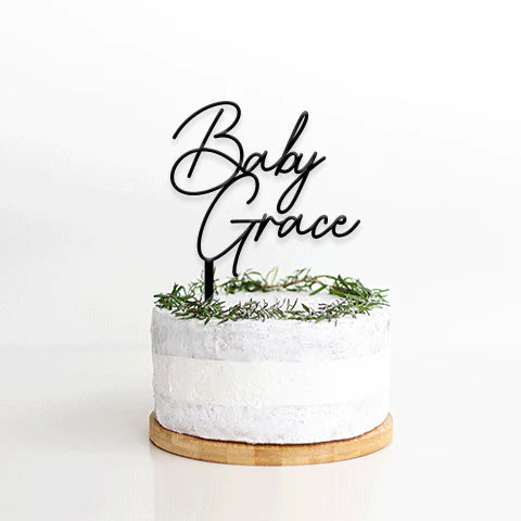 A round cake with a smooth, light-colored frosting is placed on a wooden cake stand. Atop the cake sits a customized acrylic cake topper featuring the words "Baby Grace" in an elegant, cursive font. The base of the topper is adorned with a decorative ring of green foliage. The background is plain and white, emphasizing the cake and topper.