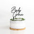 A round cake with a smooth, light-colored frosting is placed on a wooden cake stand. Atop the cake sits a customized acrylic cake topper featuring the words "Baby Grace" in an elegant, cursive font. The base of the topper is adorned with a decorative ring of green foliage. The background is plain and white, emphasizing the cake and topper.