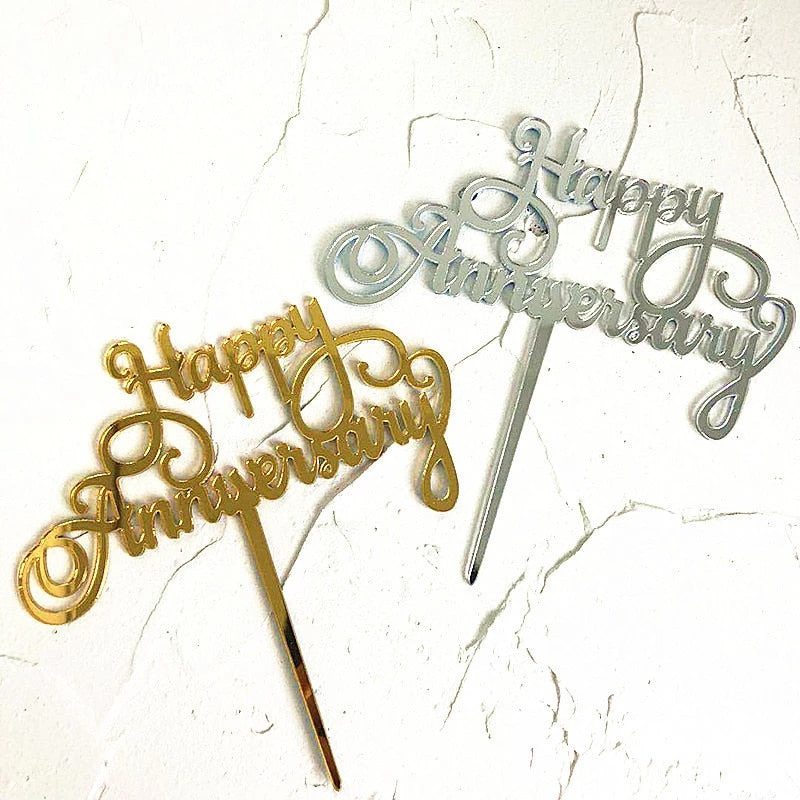 Two acrylic cake toppers displayed on a textured background. One topper is gold with the text "Happy Anniversary" in an elegant cursive font, while the other is silver with the same design. Both toppers have a pointed base for inserting into a cake.