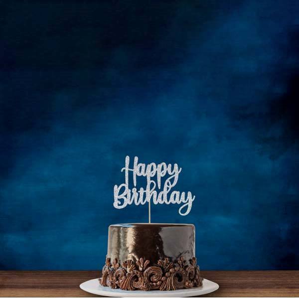 The image features a chocolate birthday cake placed on a white circular cake board. The cake has a glossy finish with decorative swirls along the bottom edge. Atop the cake, there is a silver acrylic cake topper that reads "Happy Birthday" in an elegant font. The background is a deep blue, providing a contrasting backdrop to the cake.