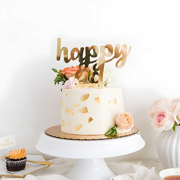 A decorative cake on a white pedestal, featuring a smooth, pale-colored frosting with gold leaf accents. Atop the cake is a golden acrylic topper that reads "happy eid" in a playful script. Surrounding the cake are fresh flowers in soft pink and green hues. In the foreground, there are additional desserts, including a cupcake with orange frosting, and a small card that says "Eid Mubarak." The background is simple and light, enhancing the festive presentation.