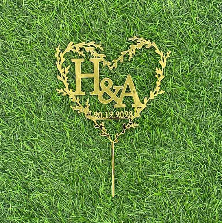 A gold acrylic cake topper in the shape of a heart, featuring the initials "H&A" in the center. The design includes decorative vine-like elements surrounding the heart. Below the initials, there is a date displayed as "20.12.2023". The topper is set against a green grass-like background.