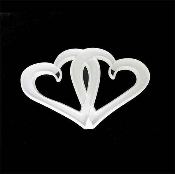 A white cookie cutter shaped like two interlocking hearts, set against a black background. The design features smooth curves and a three-dimensional appearance, highlighting the joined connection between the hearts.