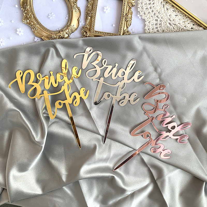 Three acrylic cake toppers displaying the text "Bride to be" in various metallic colors: gold, silver, and rose gold. The toppers are placed on a soft, folded satin fabric, with decorative picture frames in the background.
