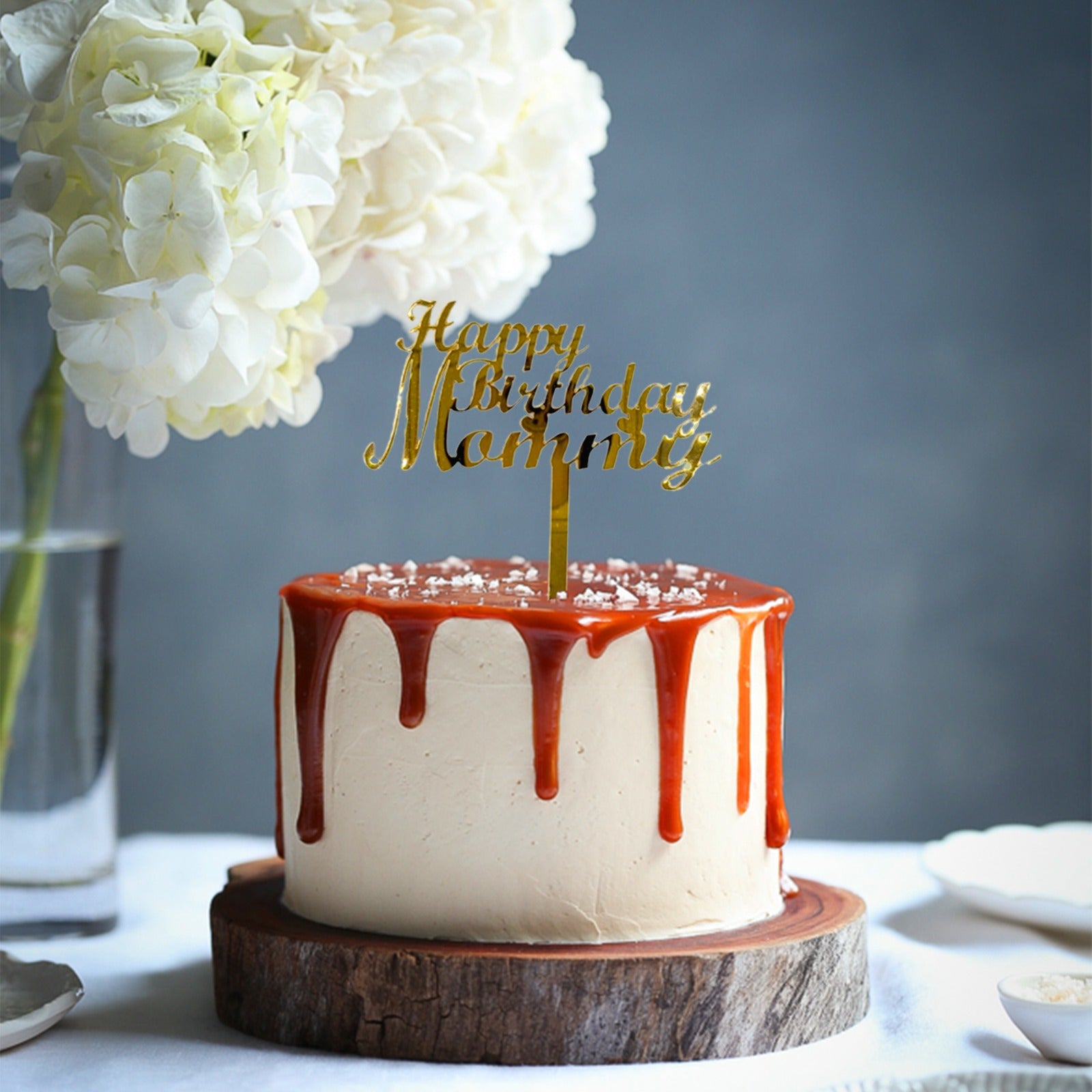 A round cake with a creamy light-colored frosting, topped with a shiny caramel drizzle that drips down the sides. An elegant acrylic topper in gold lettering reads "Happy Birthday Mommy." The cake is placed on a rustic wooden board. In the background, there is a vase with white flowers and a blurred out water glass on a light surface.