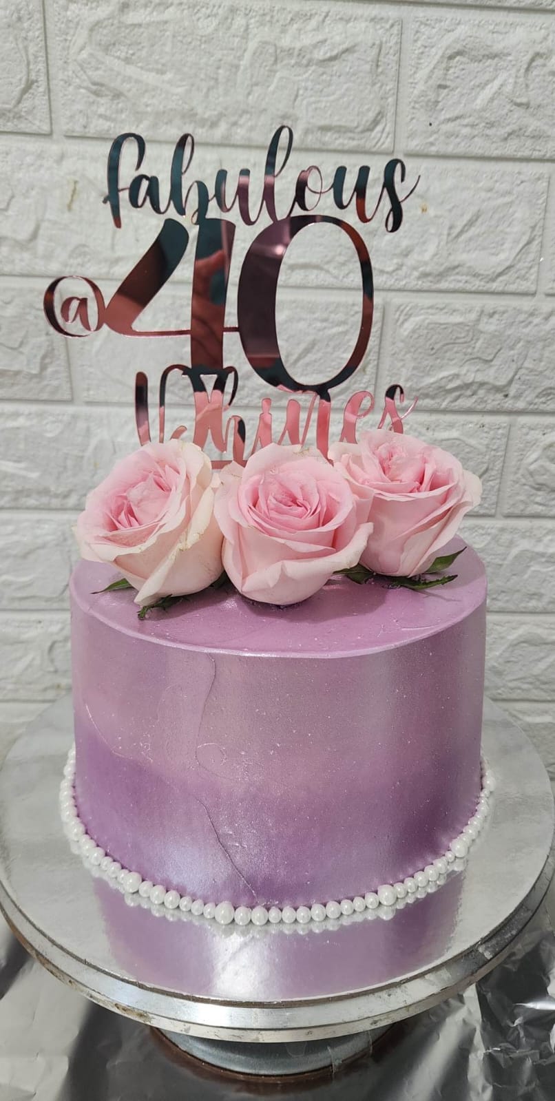 A round cake with a smooth purple ombre finish. The cake is topped with three light pink roses arranged at the center. Above the roses, there is a custom cake topper made of shiny material, reading "fabulous @ 40" in an elegant font. The cake is decorated with small white beads around the base. The background features a textured white wall.