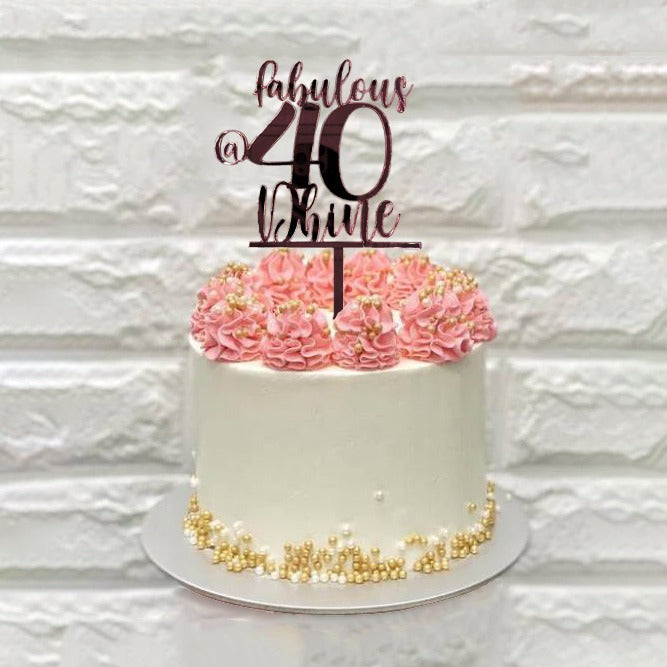 A white birthday cake with pink frosting swirls on top and decorated with small golden beads. It features a customized cake topper that reads "fabulous @ 40" in elegant script. The background has a textured white wall.