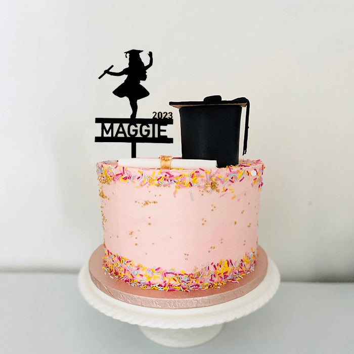 A round cake with pink frosting, decorated with colorful sprinkles around the bottom edge. Atop the cake is a black acrylic cake topper featuring a silhouette of a girl wearing a graduation cap and holding a diploma, along with the name "Maggie" and the year "2023." The cake sits on a white decorative cake stand.