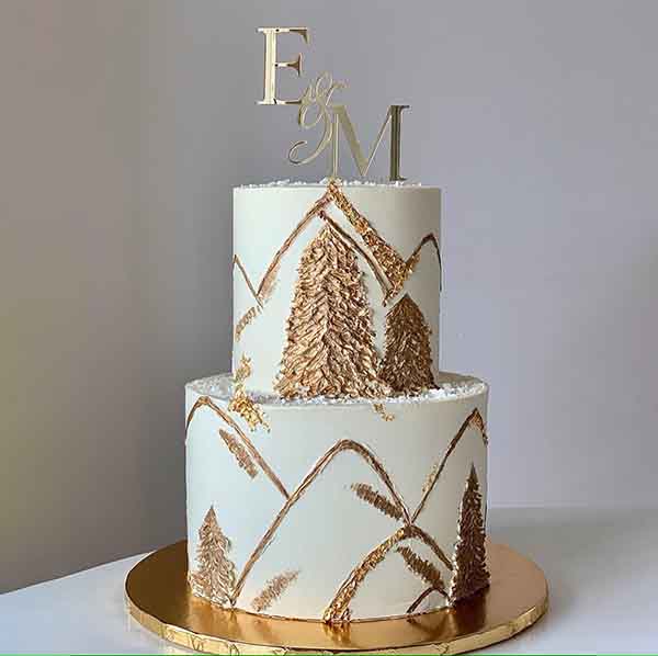 A two-tiered wedding cake with a white frosting base. The cake features gold leaf decorations and textured designs resembling trees and mountains. At the top, there is a decorative cake topper with the letters "E," "of," and "M" in elegant script. The cake is placed on a round gold cake board. The background is neutral, enhancing the cake&