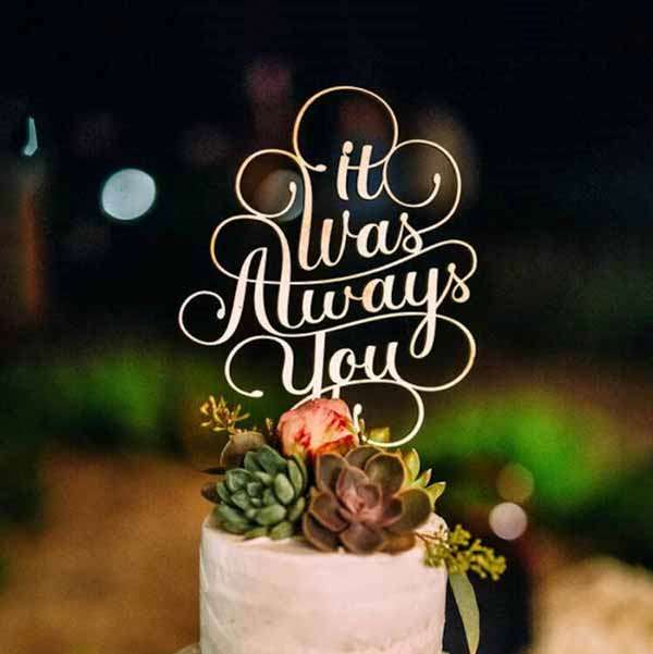 The image features a decorative cake topper that reads "It Was Always You" in an elegant, cursive font. The topper is placed atop a white cake adorned with small green succulents and a pink rose. The background is softly lit, suggesting an outdoor evening setting. Overall, the scene conveys a romantic atmosphere suitable for a wedding or celebration.